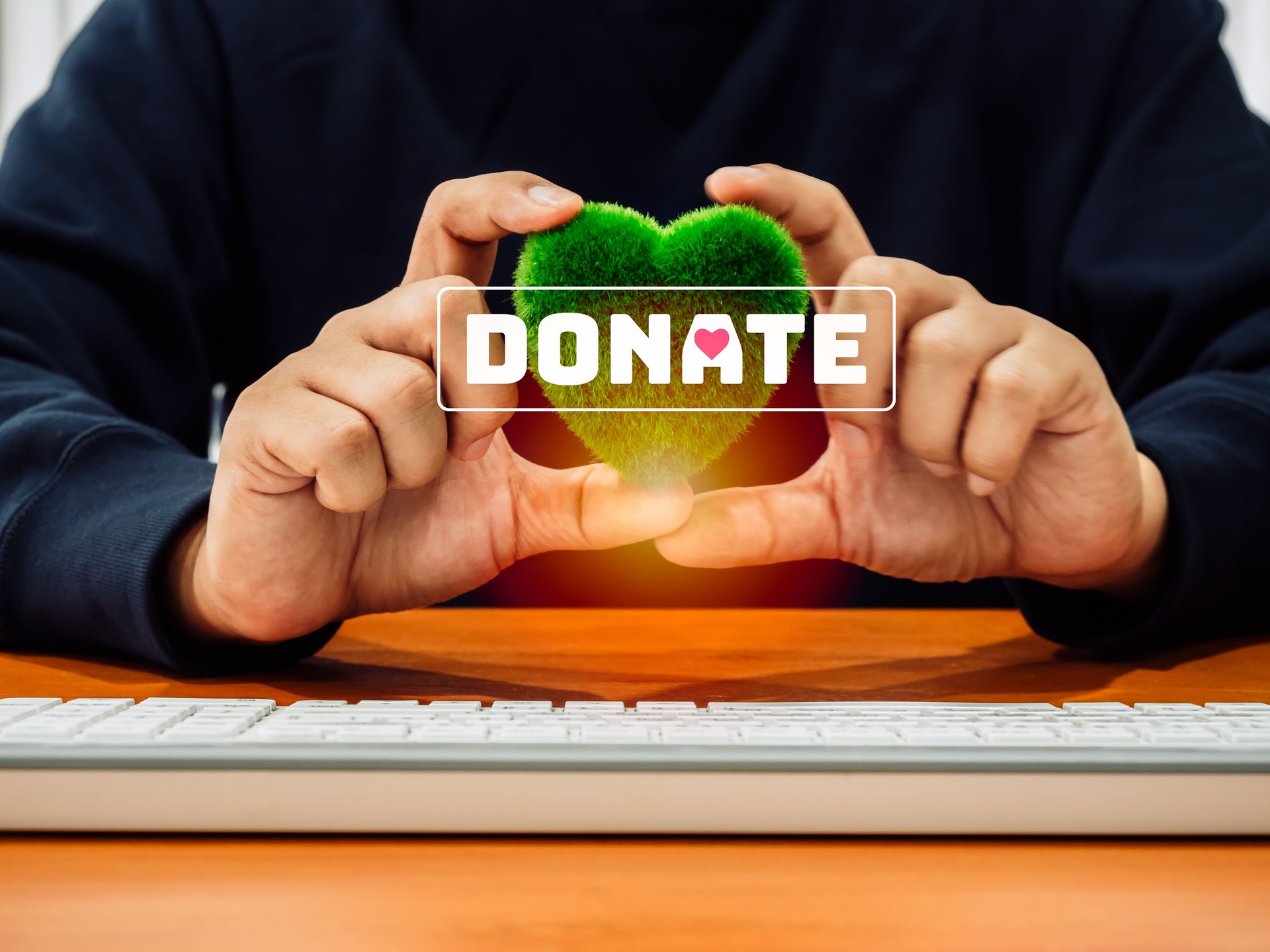 Donation online concept. Text Donate on green grass heart shape, holding by businessperson's hands who working with computer on wooden desk at workplace. Fundraising, charity donate.